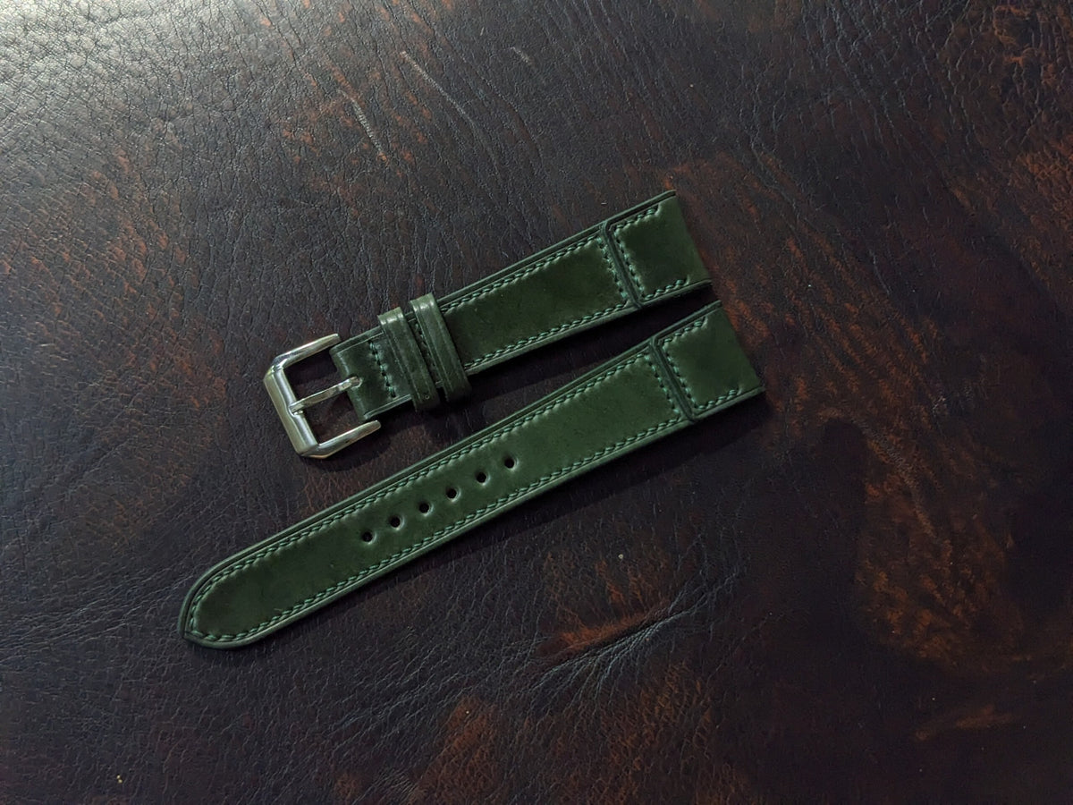 An olive green leather watch strap with a silver-toned metal buckle, laid flat on a dark brown wooden surface.