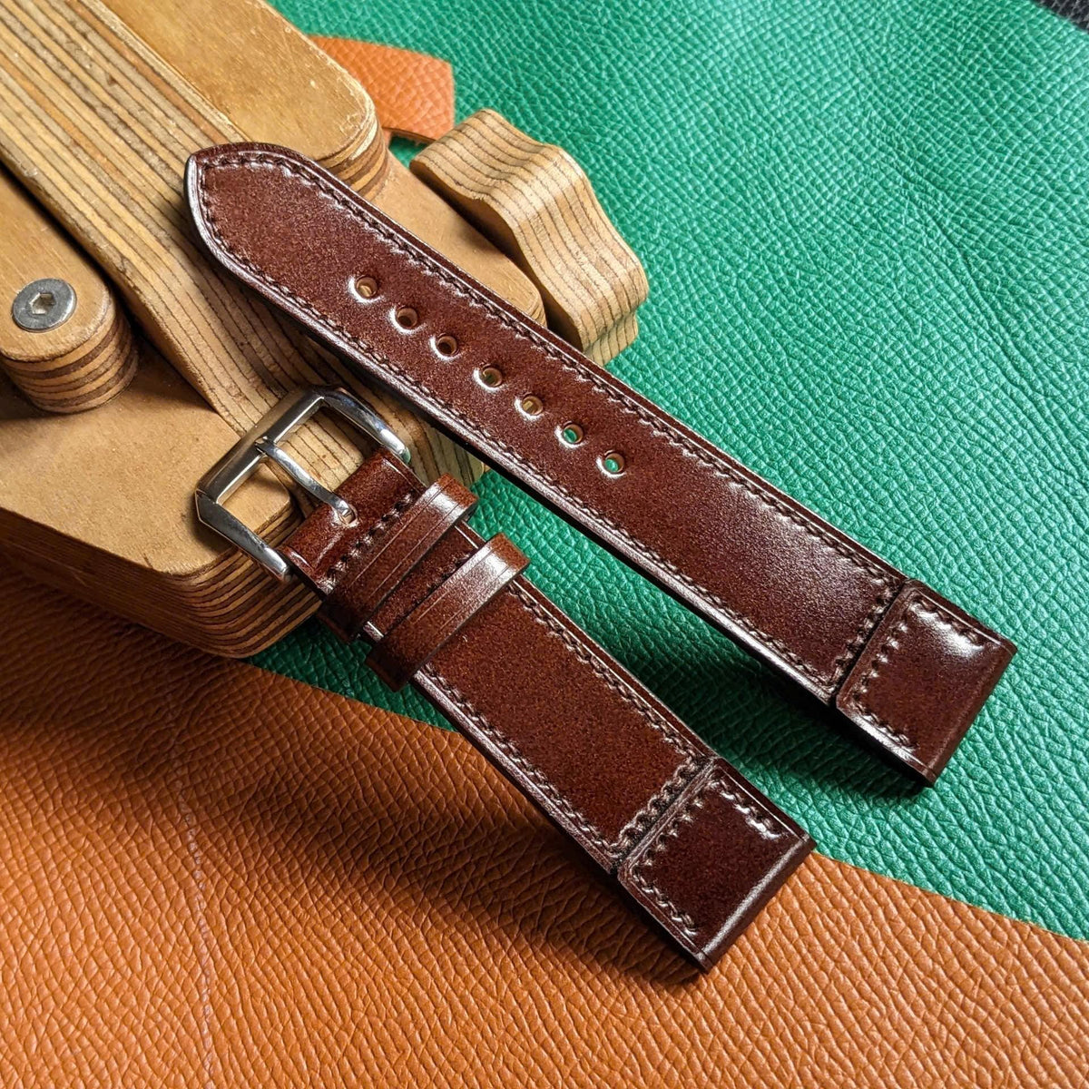 A dark brown leather watch strap with silver-tone metal hardware, laid diagonally across a green and orange textured background next to a wooden watch stand.
