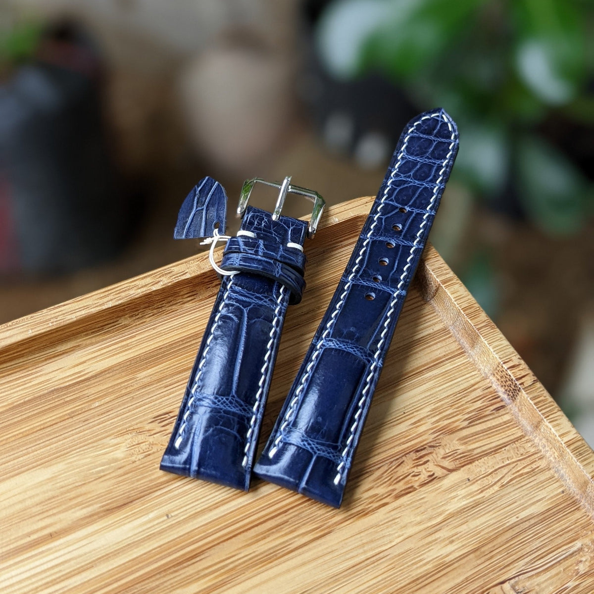 A dark blue alligator leather watch strap with white contrast stitching and a silver buckle, displayed on a light wood tray with a blurred background.