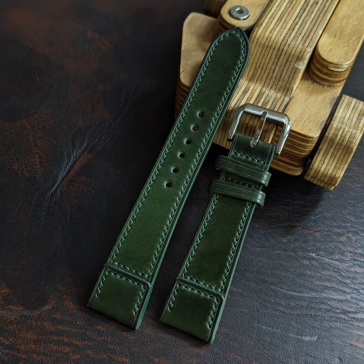 A dark green leather watch strap with a silver buckle, laid flat on a dark wooden surface next to a stack of wooden watch dials.