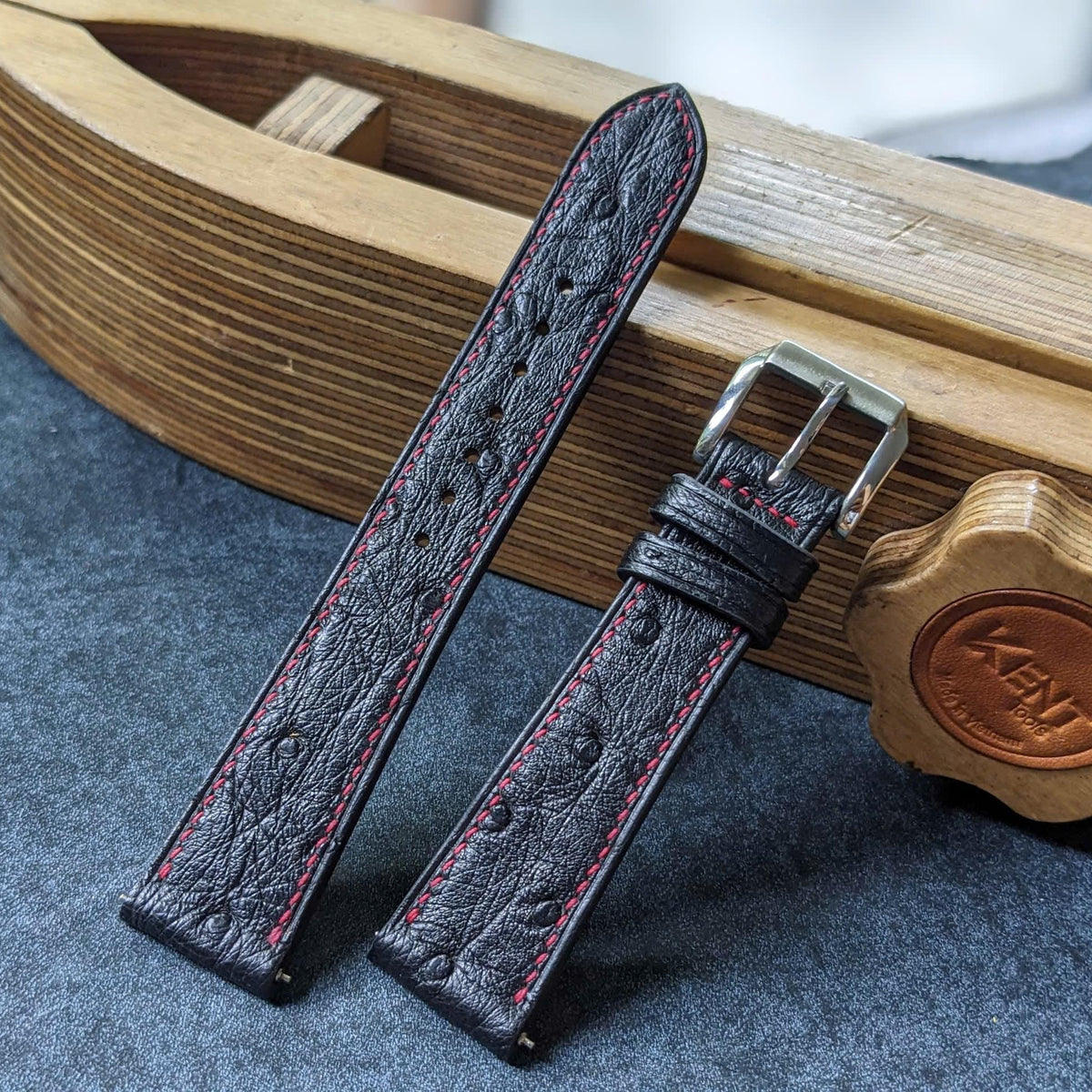 A dark brown textured leather watch strap with red stitching and a silver buckle, displayed on a gray surface next to a wooden boat-shaped box.