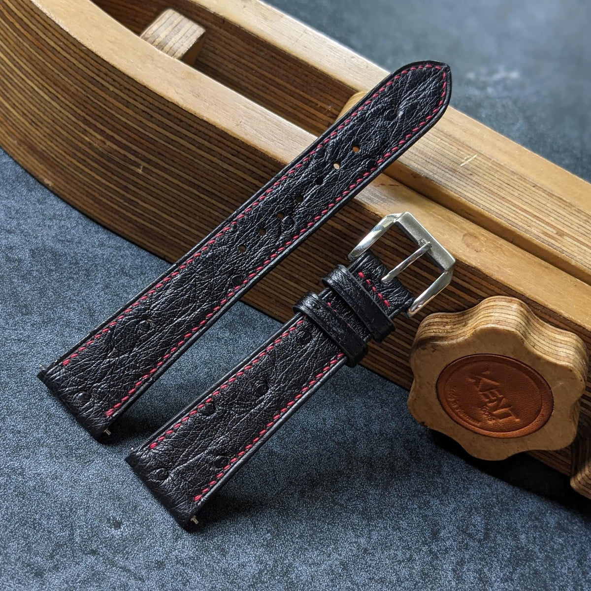 A dark brown textured leather watch strap with red stitching and a silver buckle, displayed on a gray textured surface next to a wooden watch stand.