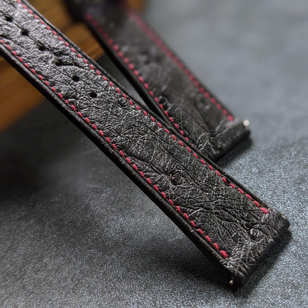 Close-up of a dark textured leather watch strap with red contrast stitching, resting on a gray textured surface.